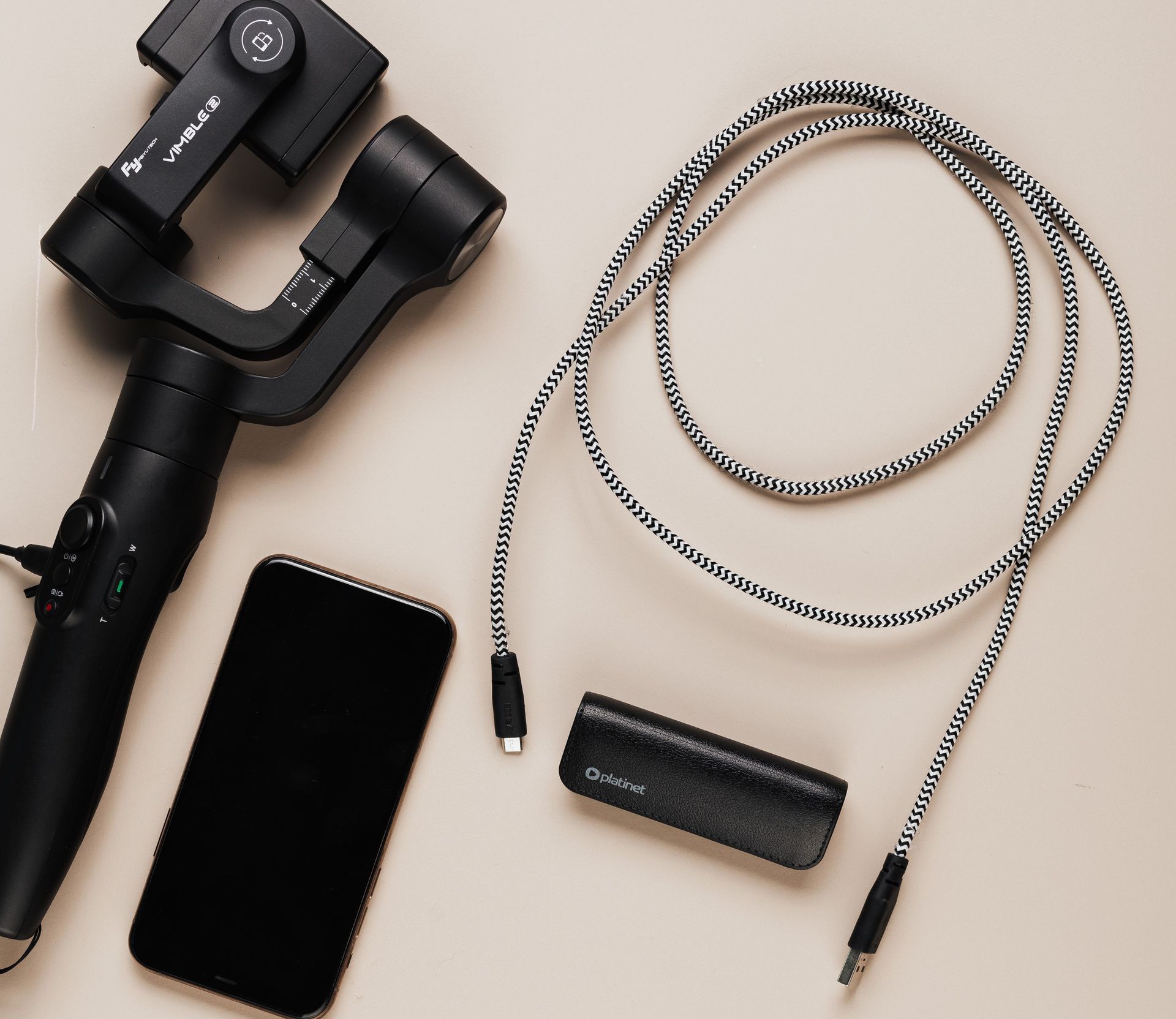 Minimalist flat lay featuring a smartphone, gimbal, charging cables, and power bank on a beige background.