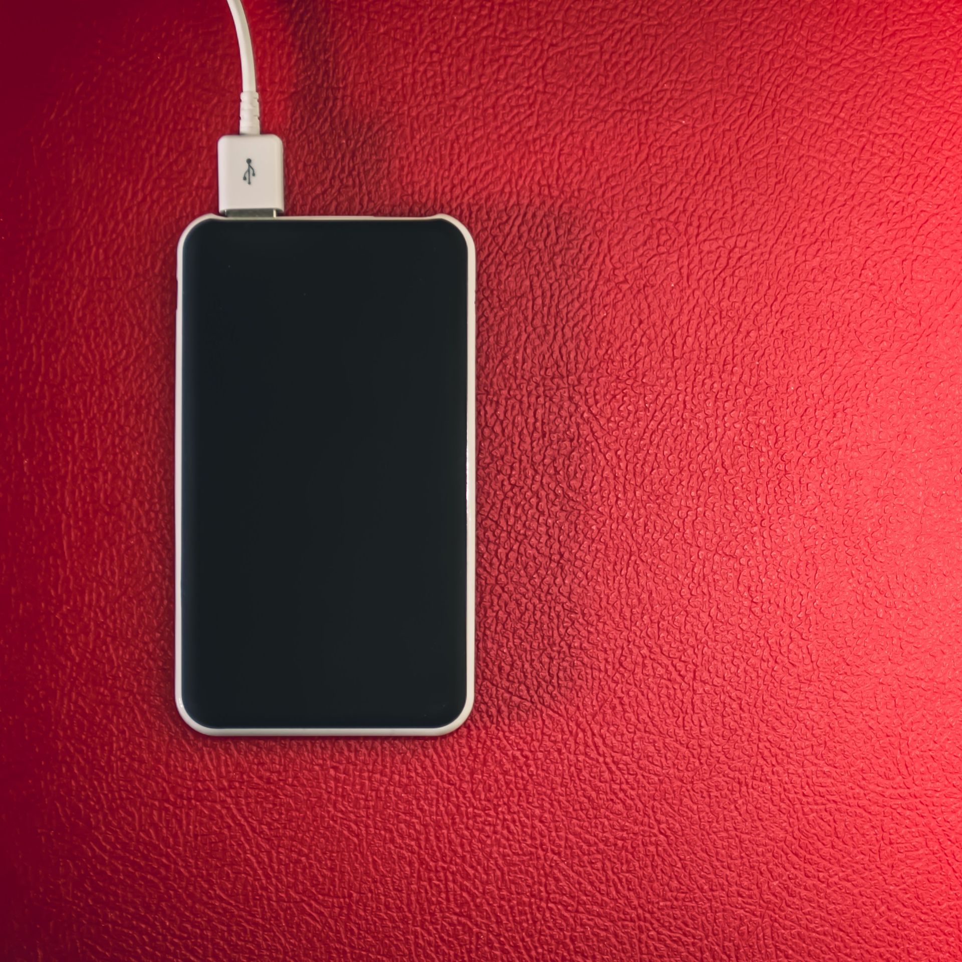 A smartphone connected to a charger placed on a red leather surface.